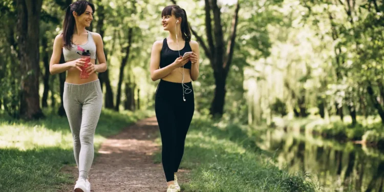 Duas mulheres caminhando em um parque verde, sorrindo e conversando durante uma caminhada ao ar livre.