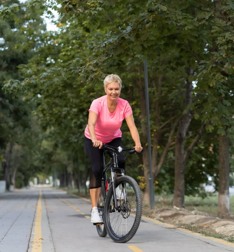 Mulher madura pedalando ao ar livre em pista cercada de árvores, com roupa esportiva.
