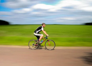 Homem pedalando em alta velocidade em estrada rural com campos verdes ao fundo, simbolizando saúde e emagrecimento através do ciclismo.