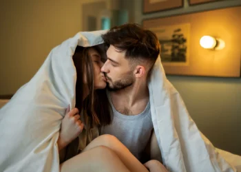 Casal apaixonado se beijando sob o edredom, curtindo um momento íntimo e especial.