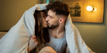 Casal apaixonado se beijando sob o edredom, curtindo um momento íntimo e especial.