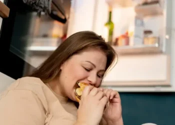 Mulher comendo compulsivamente sentada em frente à geladeira aberta.