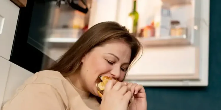 Mulher comendo compulsivamente sentada em frente à geladeira aberta.