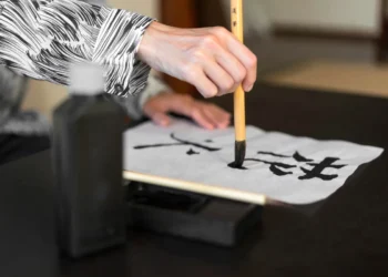 Pessoa praticando caligrafia japonesa, escrevendo a palavra "Kaizen" em papel branco com pincel de bambu.