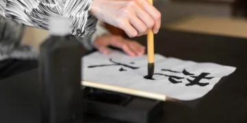 Pessoa praticando caligrafia japonesa, escrevendo a palavra "Kaizen" em papel branco com pincel de bambu.