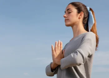 Mulher meditando ao ar livre com as mãos em posição de oração, com expressão tranquila, respirando para aliviar o estresse.