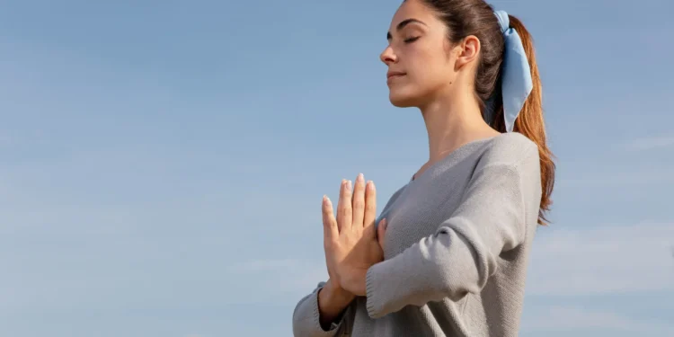 Mulher meditando ao ar livre com as mãos em posição de oração, com expressão tranquila, respirando para aliviar o estresse.