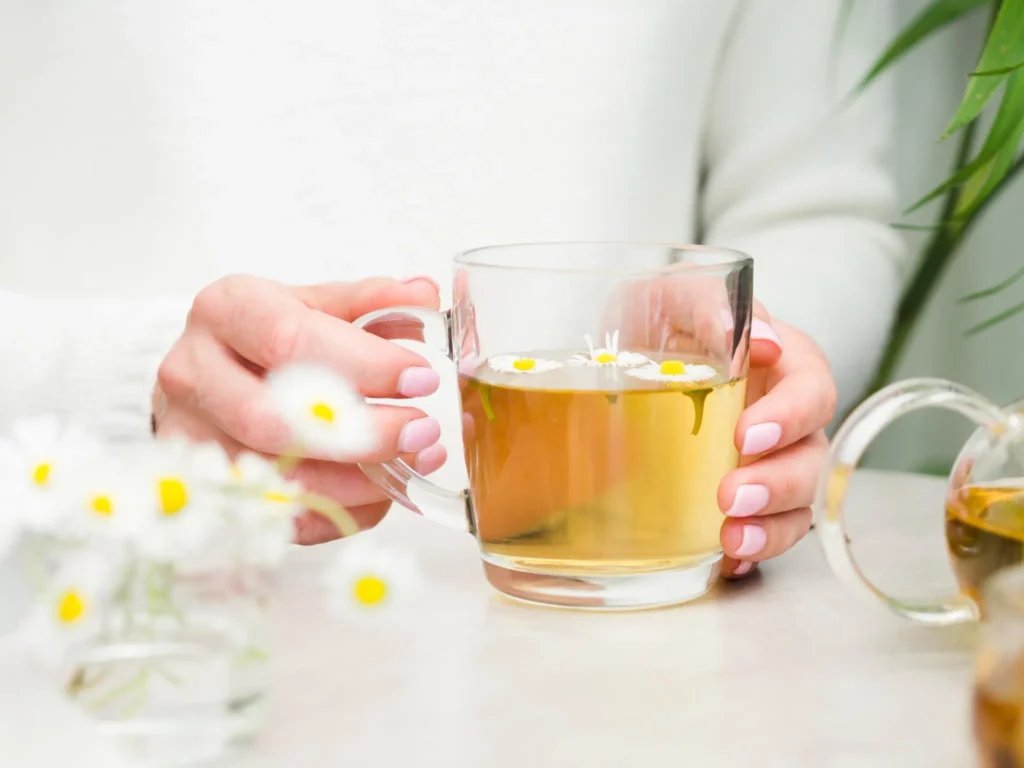 Mãos segurando uma xícara de chá de camomila com flores flutuando, representando técnicas naturais para melhorar o sono.