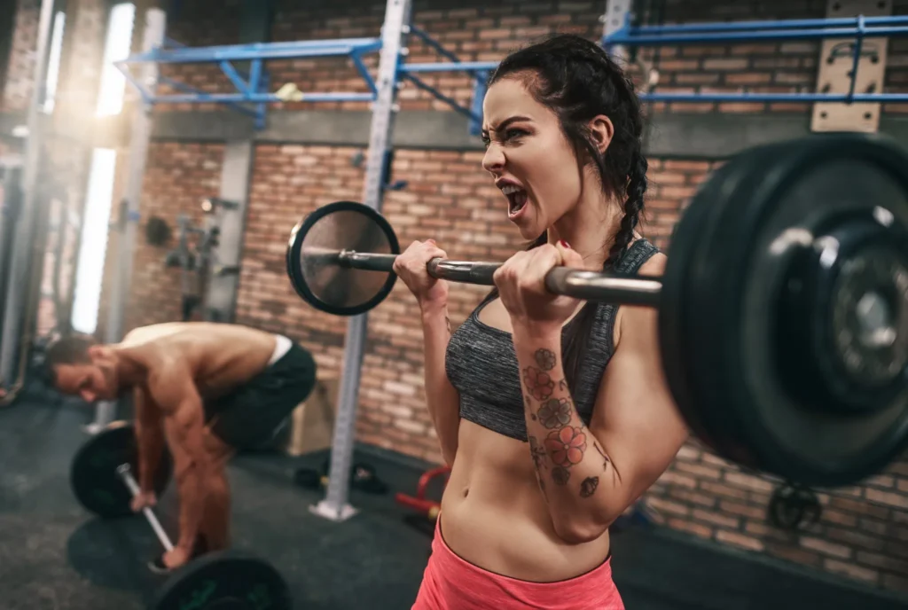 Mulher levantando halteres com foco no treino de força, enquanto homem treina ao fundo na academia.