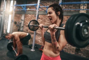 Mulher levantando halteres com foco no treino de força, enquanto homem treina ao fundo na academia.