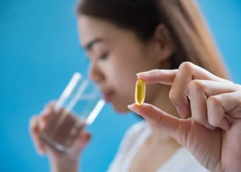 Mulher segurando cápsula de vitamina B12 enquanto bebe água, fundo azul desfocado