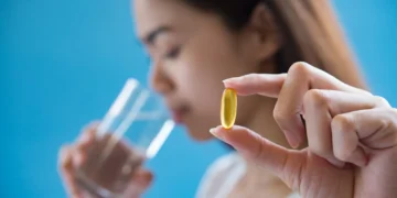 Mulher segurando cápsula de vitamina B12 enquanto bebe água, fundo azul desfocado