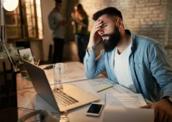 Homem exausto tentando se concentrar em ambiente de trabalho barulhento e desorganizado.