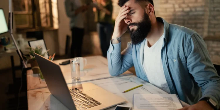 Homem exausto tentando se concentrar em ambiente de trabalho barulhento e desorganizado.