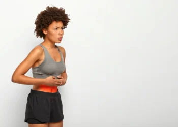 Mulher afro-americana com dor abdominal, tocando a barriga e mostrando expressão de desconforto, usando top esportivo cinza e shorts preto, com fundo branco.