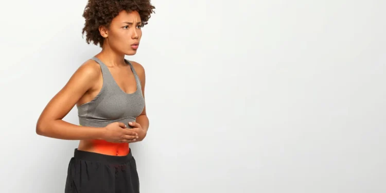 Mulher afro-americana com dor abdominal, tocando a barriga e mostrando expressão de desconforto, usando top esportivo cinza e shorts preto, com fundo branco.