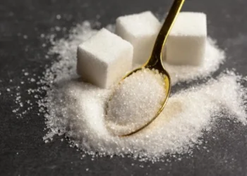 Cubos de açúcar e grânulos dispersos sobre uma superfície escura, com uma colher dourada cheia de açúcar, ilustrando os efeitos de parar de consumir açúcar.