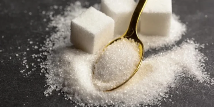 Cubos de açúcar e grânulos dispersos sobre uma superfície escura, com uma colher dourada cheia de açúcar, ilustrando os efeitos de parar de consumir açúcar.