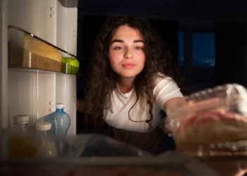 Mulher abrindo a geladeira à noite, com um olhar atento e curiosidade.