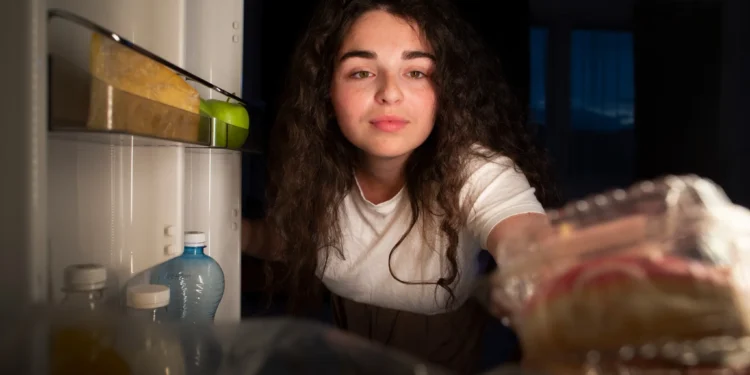 Mulher abrindo a geladeira à noite, com um olhar atento e curiosidade.