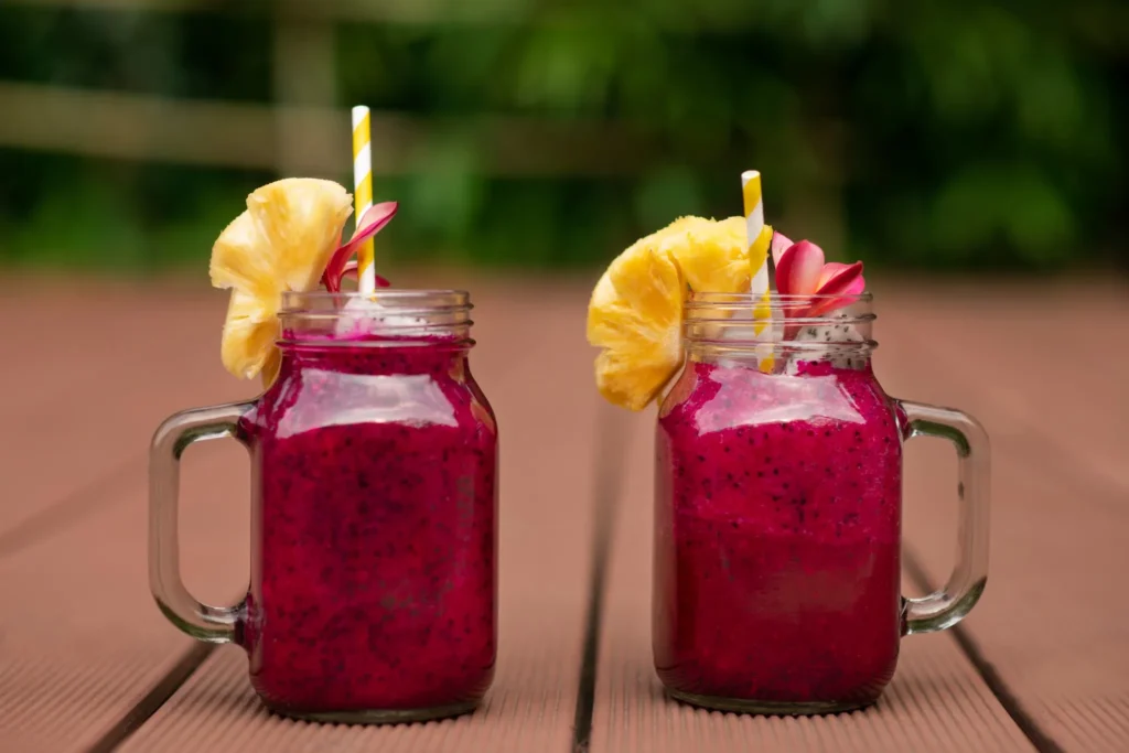 Dois copos de vidro com smoothies roxos decorados com abacaxi e flores tropicais