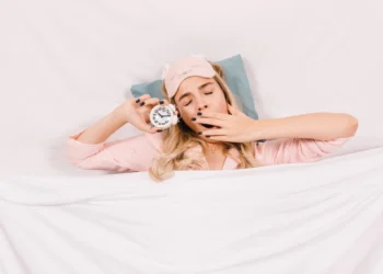 Mulher sonolenta na cama com despertador, representando a importância do sono para o bem-estar