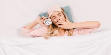 Mulher sonolenta na cama com despertador, representando a importância do sono para o bem-estar