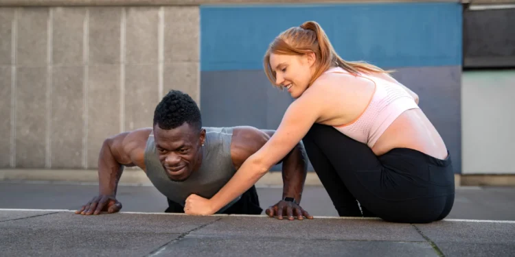 Homem fazendo flexão ao ar livre com a ajuda de uma mulher durante treino para ganho de músculos e definição corporal.