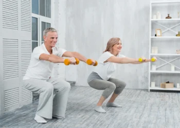 Casal mais velho realizando agachamento com halteres em casa, em uma atividade de fortalecimento muscular segura após os 60 anos.
