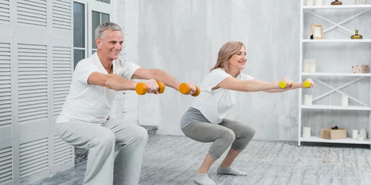 Casal mais velho realizando agachamento com halteres em casa, em uma atividade de fortalecimento muscular segura após os 60 anos.