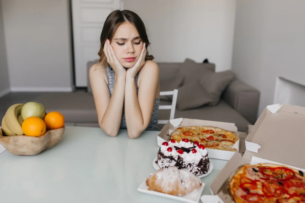 Mulher olhando para uma mesa com pizza e doces enquanto tenta resistir à tentação durante uma dieta, refletindo sobre a armadilha do prazer instantâneo dos alimentos ultraprocessados.