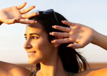 Mulher ao ar livre, sorrindo, cobrindo os olhos com a mão para se proteger do sol, representando a importância da exposição solar para a produção de vitamina D.