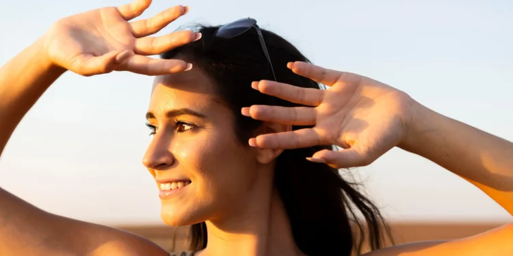 Mulher ao ar livre, sorrindo, cobrindo os olhos com a mão para se proteger do sol, representando a importância da exposição solar para a produção de vitamina D.