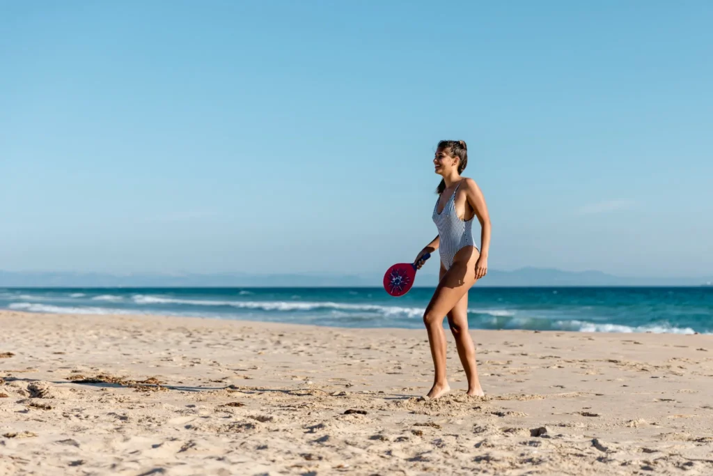 Mulher jogando beach tennis na praia, com sorriso e energia positiva.