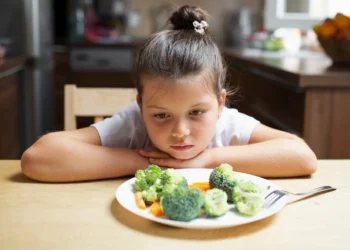 Criança olhando com desânimo para um prato com legumes e verduras em uma cozinha.