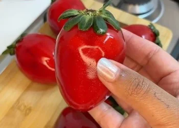 Imagem de um morango do amor grande e brilhante, sendo segurado por uma mão, com outras frutas ao fundo.