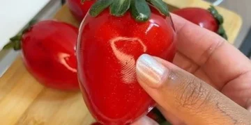 Imagem de um morango do amor grande e brilhante, sendo segurado por uma mão, com outras frutas ao fundo.