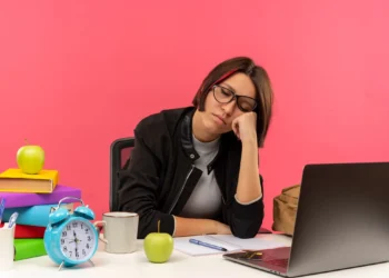 Mulher com óculos, sonolenta e com a mão na bochecha, diante de uma mesa cheia de material de estudo, mostrando a exaustão de uma rotina intensa.