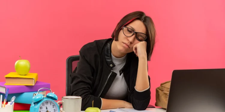 Mulher com óculos, sonolenta e com a mão na bochecha, diante de uma mesa cheia de material de estudo, mostrando a exaustão de uma rotina intensa.