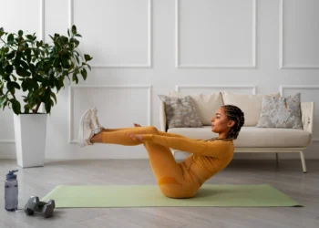 Mulher realizando exercício fitness em casa com postura de barco em tapete de yoga, com roupas de ginástica douradas e tênis branco, em ambiente interno com plantas e sofá.
