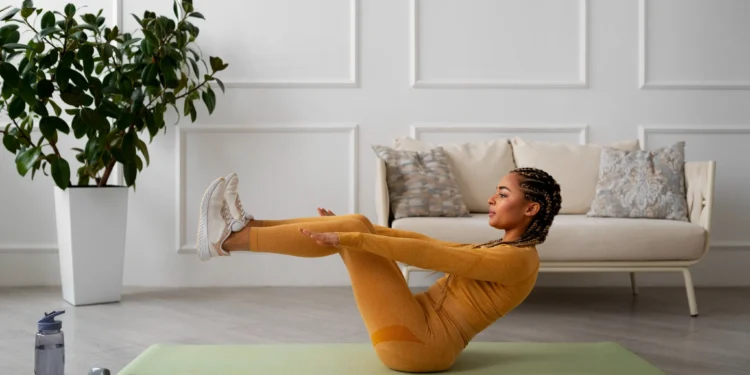 Mulher realizando exercício fitness em casa com postura de barco em tapete de yoga, com roupas de ginástica douradas e tênis branco, em ambiente interno com plantas e sofá.