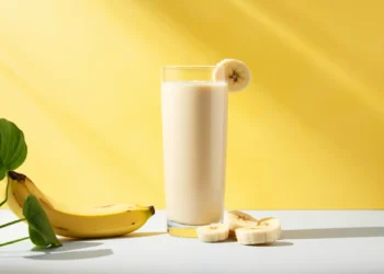 Shake de banana cremoso em copo alto com fatias de banana e uma banana inteira ao lado, em fundo amarelo.