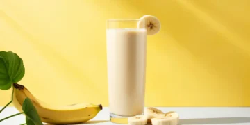 Shake de banana cremoso em copo alto com fatias de banana e uma banana inteira ao lado, em fundo amarelo.