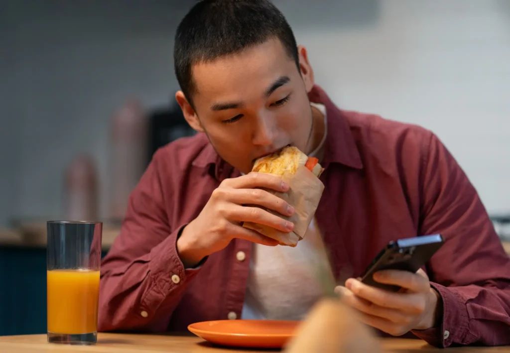 Homem comendo sanduíche enquanto segura o celular, simbolizando como a falta de tempo e distrações afetam nossos hábitos alimentares.