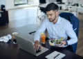 Homem jovem trabalhando no laptop enquanto almoça no escritório, com um prato de salada e uma bebida ao lado.