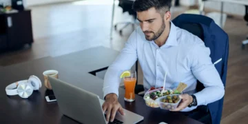 Homem jovem trabalhando no laptop enquanto almoça no escritório, com um prato de salada e uma bebida ao lado.