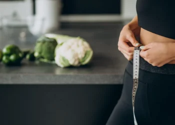 Mulher ajustando fita métrica ao redor da cintura na cozinha, com vegetais frescos ao fundo, simbolizando o processo de emagrecimento com alimentação simples.