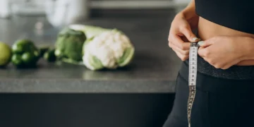 Mulher ajustando fita métrica ao redor da cintura na cozinha, com vegetais frescos ao fundo, simbolizando o processo de emagrecimento com alimentação simples.