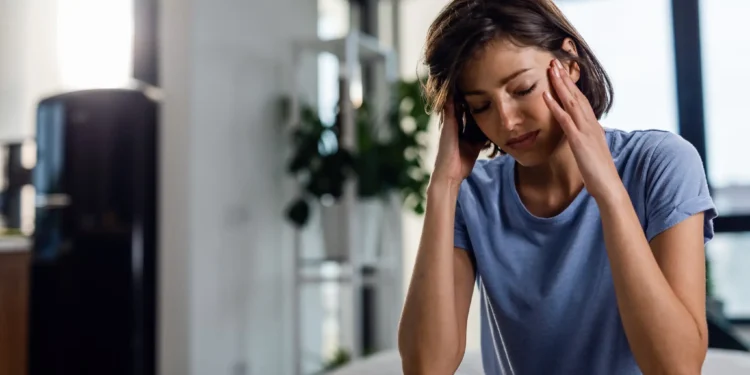 Mulher com expressão de dor, segurando a cabeça em um quarto, sugerindo sinais de exaustão emocional.