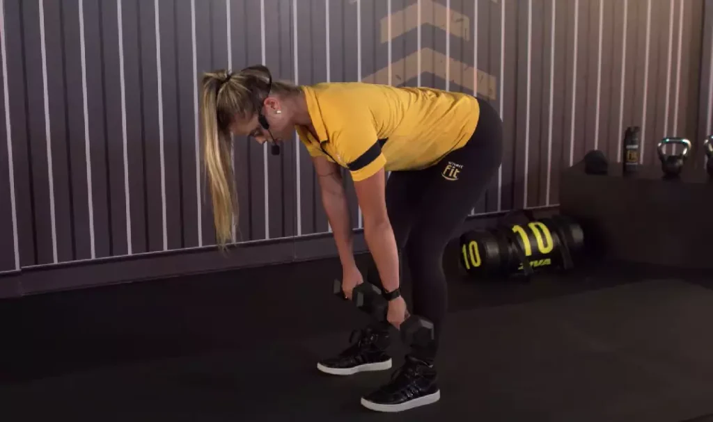 Mulher realizando exercício de stiff com halteres na academia, focando no alongamento dos glúteos e posteriores.